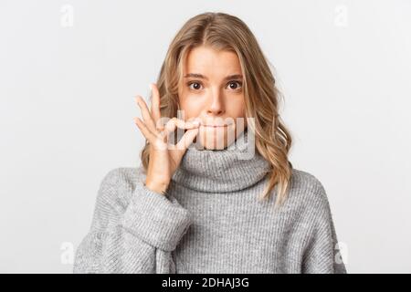 Nahaufnahme von niedlichen blonden Mädchen in grauen Pullover, halten ihr Versprechen, Zippen Lippen, Speichern Geheimnis, stehen über weißem Hintergrund Stockfoto