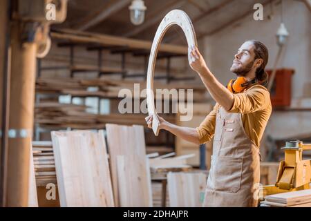 Portrait eines schönen Zimmermanns, der auf das Holzprodukt schaut und in der Werkstatt Waren mit Sperrholz baut Stockfoto