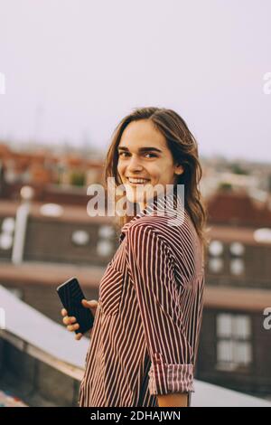 Porträt einer glücklichen jungen Frau, die das Mobiltelefon im Stehen hält Terrasse in der Stadt Stockfoto
