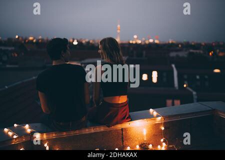 Rückansicht des jungen Paares, das auf der beleuchteten Terrasse sitzt Stadt in der Abenddämmerung Stockfoto