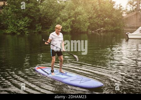 Volle Länge des Senior man Paddleboarding auf See während SUP Kurs Stockfoto