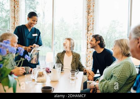 Lächelnde Krankenschwester, die der älteren Frau, die mit sitzt, einen Drink serviert Freunde und Enkel am Esstisch im Pflegeheim Stockfoto