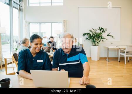 Lächelnd junge weibliche Gesundheitsarbeiterin, die älteren Mann bei der Verwendung unterstützt Laptop am Esstisch Stockfoto