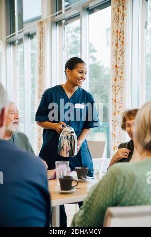 Lächelnde Krankenschwester, die älteren Frauen und Männern Getränke serviert Am Esstisch im Pflegeheim Stockfoto