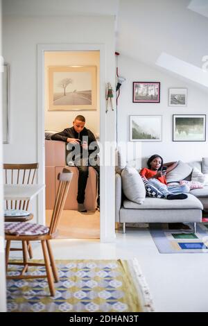 Mutter benutzt Mobiltelefon auf dem Sofa, während Junge im Sofa sitzt Abstellraum zu Hause Stockfoto