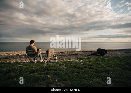 Volle Länge der Mann mit Laptop und Handy, während Sitzen am Strand gegen bewölkten Himmel Stockfoto