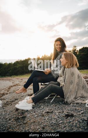 Lächelnde Frau mit Laptop, während sie mit einem Freund am Strand sitzt Bei kaltem Wetter Stockfoto