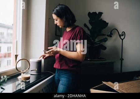Frau hält Smartphone, während sie Geräte am Fenster benutzt Stockfoto