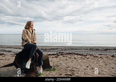 Fröhliche junge Frau, die am Strand auf verwittertem Metall sitzt Himmel Stockfoto