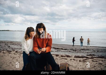 Ein Paar schaut auf das Handy, während es am Meer sitzt Am Strand Stockfoto