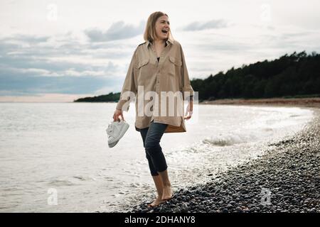 Fröhliche junge Frau, die Schuhe hält, während sie am Strand dagegen läuft Himmel Stockfoto