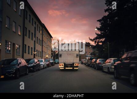 Lieferwagen bewegt sich auf der Straße zwischen geparkten Autos in der Stadt Gegen den Himmel bei Sonnenuntergang Stockfoto