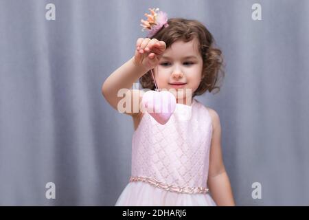 Ein rosa weiches Spielzeug in Form eines Herzens in den Händen eines 4-5-jährigen Mädchens. Valentinstag zum Valentinstag. Nahaufnahme im Hochformat auf grauem Hintergrund Stockfoto