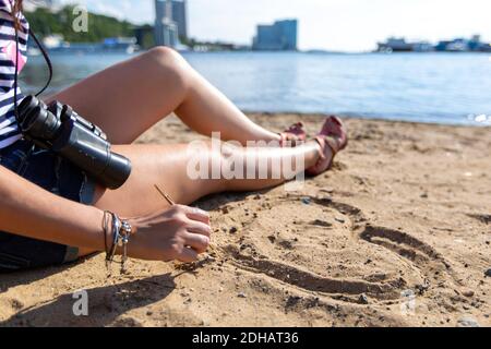 Ein junges Mädchen in einer Weste mit Fernglas sitzt auf dem Sand in der Nähe des Meeres in der Stadt und zeichnet ein Herz mit einem Stock. Stockfoto