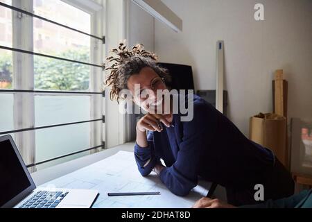 Lächelnde junge Architektin, die sich im Büro auf den Tisch lehnt Stockfoto