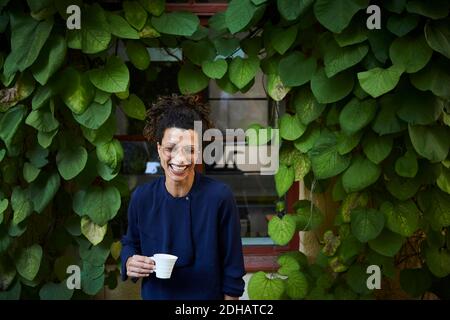 Glückliche junge Architektin hält Kaffeetasse gegen kriechenden Pflanzen Im Hinterhof Stockfoto