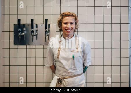 Porträt einer lächelnden Küchenchefin, die an der Wand mit weißen Kacheln steht In der Restaurantküche Stockfoto