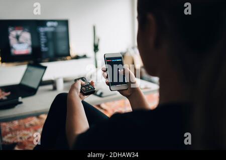 Rückansicht des Teenagers mit Handy-App und Fernbedienung Steuern Sie, während Sie Smart TV zu Hause ansehen Stockfoto