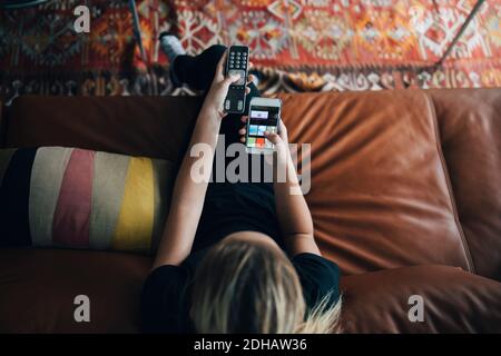 High-Angle-Ansicht von Teenager-Mädchen mit Telefon-App und Fernbedienung, während Sie auf dem Sofa sitzen und zu Hause Fernsehen Stockfoto