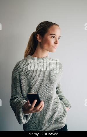 Teenager-Mädchen hält Handy, während sie gegen weiße Wand stehen Zu Hause Stockfoto