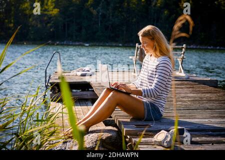 Frau mit Kopfhörern mit Laptop, während sie auf dem Steg sitzt see Stockfoto