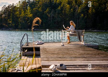 Mittlere Erwachsene Frau macht Videoanruf auf Laptop, während sie sitzt Auf Bank über Holzsteg gegen See im Wald Stockfoto