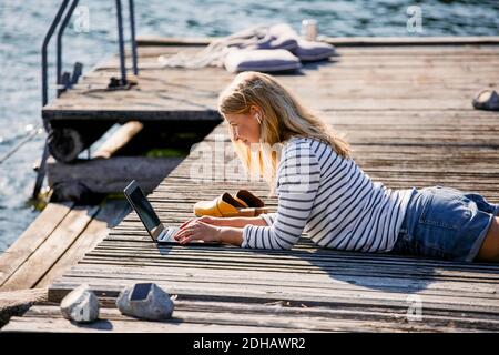 Mittlere Erwachsene Frau, die auf dem Laptop tippt, während sie über Holz liegt pier während Sommerferien Stockfoto