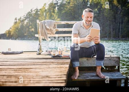 Lächelnder, reifer Mann mit digitalem Tablet und in-Ear-Kopfhörern Sitzen auf dem Steg über dem See Stockfoto