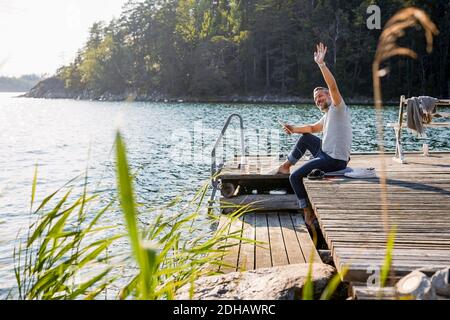 Reifer Mann, der wegschaut und winkt, während er das digitale Tablet hält Auf dem Steg über dem See Stockfoto