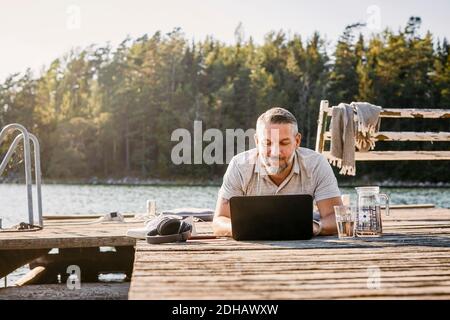 Lächelnder Mann, der auf der Vorderseite liegt, während er einen Laptop über Holz benutzt Steg Stockfoto