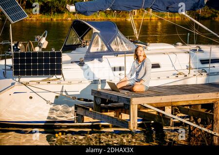 Nachdenkliche Frau mit Laptop sitzt auf dem Pier an der festgetäuten Yacht Am sonnigen Seeufer Stockfoto