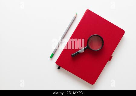 Home Office Schreibtisch mit rotem Notizbuch, Pflanze, Lupe und Bleistiften auf weißem minimalistischem Hintergrund mit Kopierfläche. Minimaler Arbeitsplatz Stockfoto