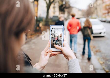 Mädchen fotografieren Freunde durch Handy, während zu Fuß auf Bürgersteig Stockfoto