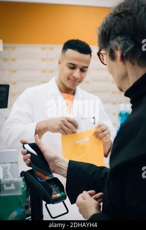 Ältere weibliche Kundin, die kontaktloses Bezahlen über das Mobiltelefon verwendet Apotheker beim Auschecken im Laden Stockfoto