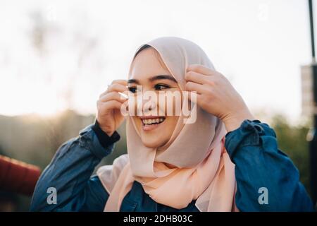 Lächelnde junge muslimische Frau, die Hijab gegen klaren Himmel anpasst Stockfoto