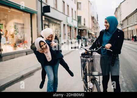Junge Frau, die mit dem Fahrrad geht und ein fröhliches Teenager-Mädchen anschaut Geben Huckepack an Freund auf der Straße in der Stadt Stockfoto