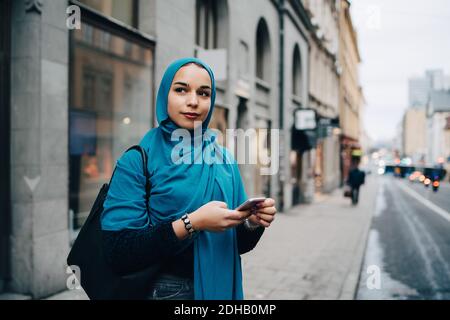 Selbstbewusste junge muslimische Frau hält Smartphone auf Bürgersteig stehen In der Stadt Stockfoto
