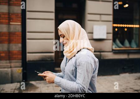 Seitenansicht einer jungen Frau, die beim Gehen Smartphone benutzt Auf der Straße in der Stadt Stockfoto