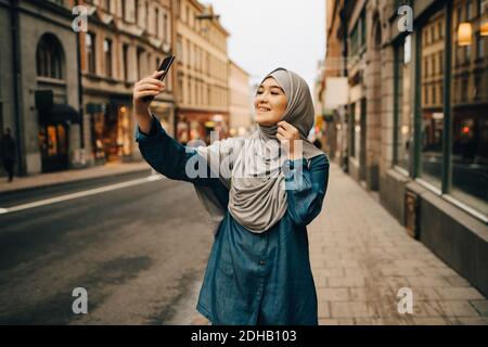 Glückliche junge muslimische Frau, die Selfie macht und auf dem Bürgersteig steht Stadt Stockfoto