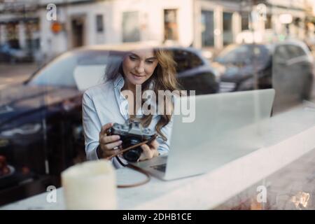 Junge Frau mit Kamera, während sie mit Laptop im Café sitzt Durch das Glasfenster gesehen Stockfoto