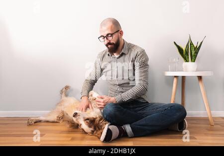 Mann stetet seinen Hund, der zu Hause auf dem Boden sitzt. Innenleben, Quarantäne, einfache Freude am Lockdown-Konzept: Tausendjährige männliche Person spielt mit seinem Stockfoto