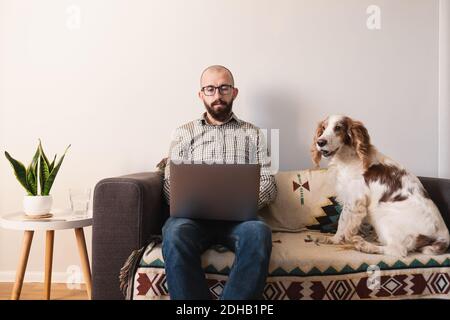 Mann arbeitet zu Hause, Hund sitzt neben ihm auf der Couch. Arbeiten aus der Ferne, Sperrung, Quarantäne Lifestyle-Konzept: Zuhause mit Haustieren mit der Arbeit kombiniert Stockfoto