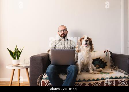 Mann arbeitet zu Hause, glücklicher Hund sitzt neben ihm auf der Couch. Arbeiten aus der Ferne, Sperrung, Quarantäne Lifestyle-Konzept: Zuhause mit Haustieren mit der kombiniert Stockfoto