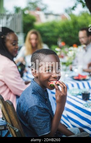 Lächelnder Junge, der Wassermelone isst, während er mit der Familie am Tisch sitzt Im Garten Stockfoto
