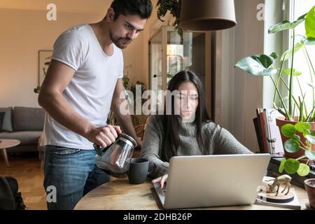 Frau mit Laptop, während Mann Gießen Kaffee in Tasse auf Tabelle Stockfoto