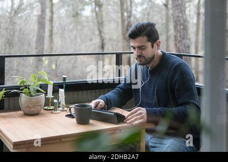 Mann mit Kopfhörern mit digitalem Tablet, während er am Tisch sitzt Auf dem Balkon Stockfoto