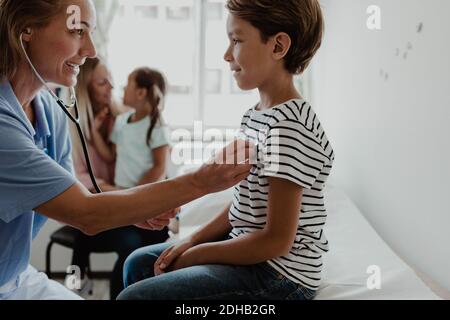 Lächelnde Ärztin, die dem Herzschlag des Jungen lauscht, während sie in der Familie ist Hintergrund in der Klinik Stockfoto