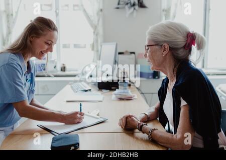 Lächelnder Arzt schreibt ein Rezept, während eine ältere Frau am Schreibtisch sitzt Im Krankenhaus Stockfoto