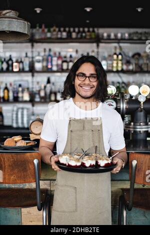 Porträt eines lächelnden Kellners, der während des Stehens Dessert im Tablett hält Gegen den Tresen im Restaurant Stockfoto