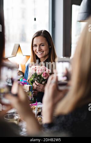 Freundinnen fotografieren lächelnde Frau mit Blumenstrauß am Esstisch Im Restaurant Stockfoto